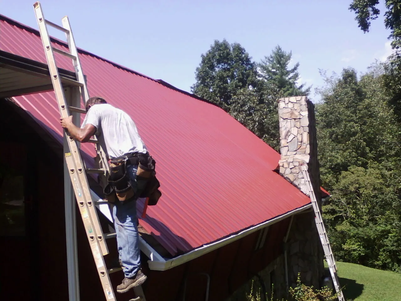 Roofer on a ladder beside a red metal roof and stone chimney — Soffit & Fascia Repair by Roofing Brothers in Lava Hot Springs